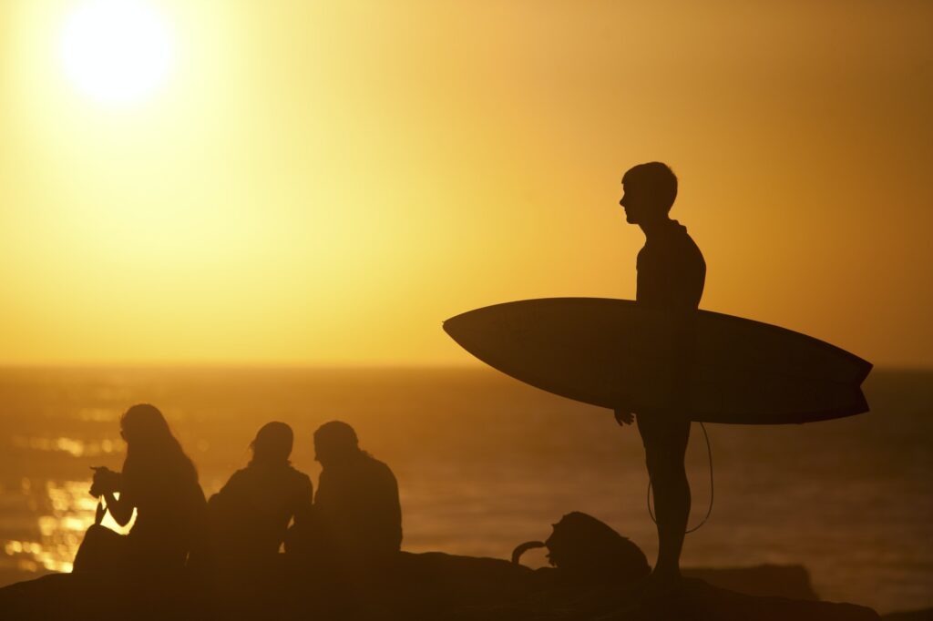 Surfer carrying surfboard on beach at sunset, Taghazout, Morocco